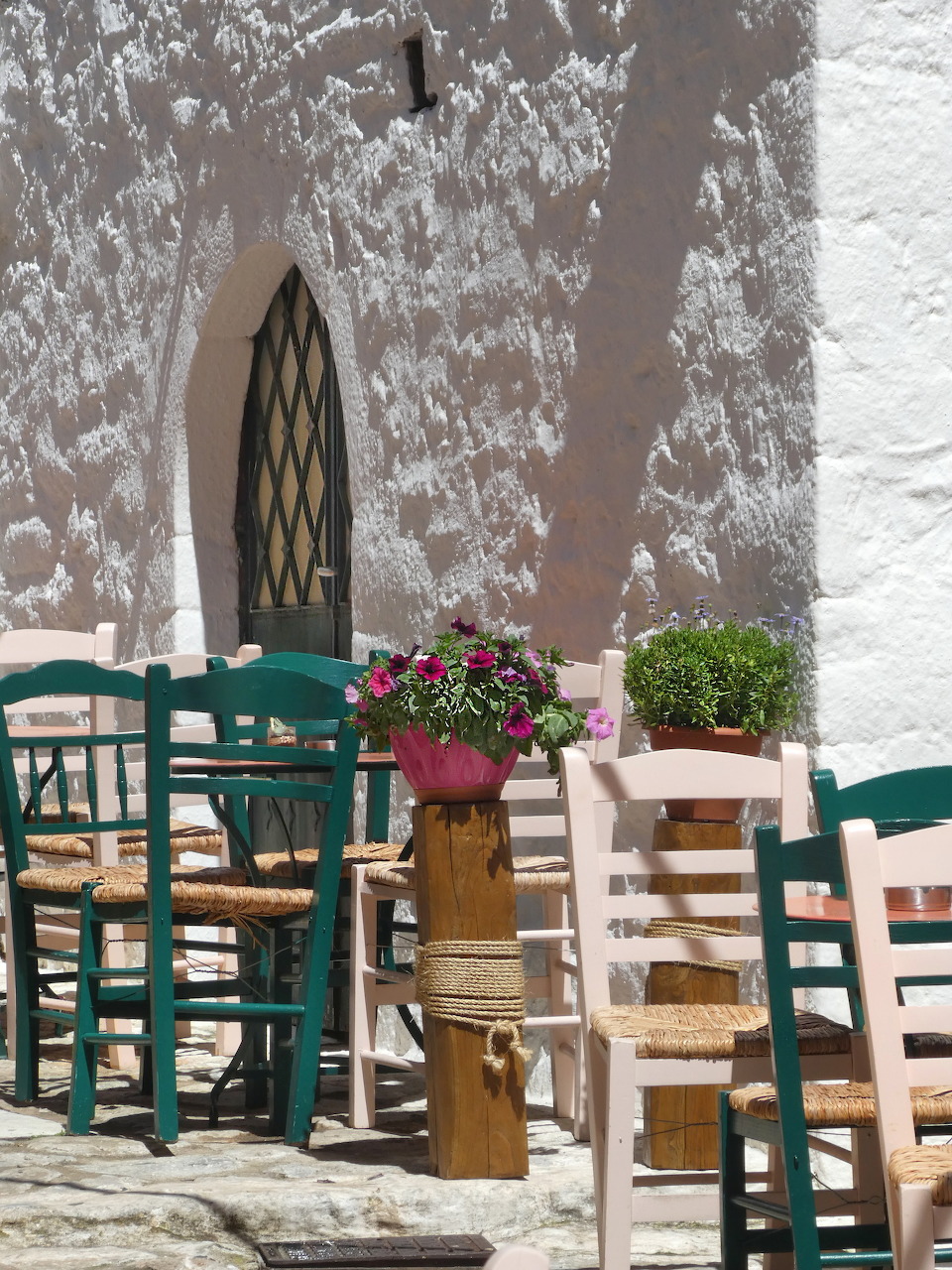 Wooden café chairs and tables with red geraniums against weathered white wall in Greek village