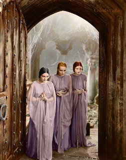 Three women in long purple dresses standing in a gothic stone archway with cobwebs and ruins