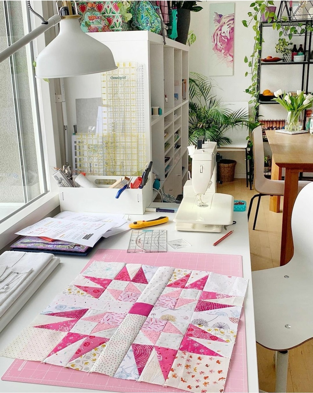 Pink star quilt block on a craft desk in a bright sewing room with plants and supplies