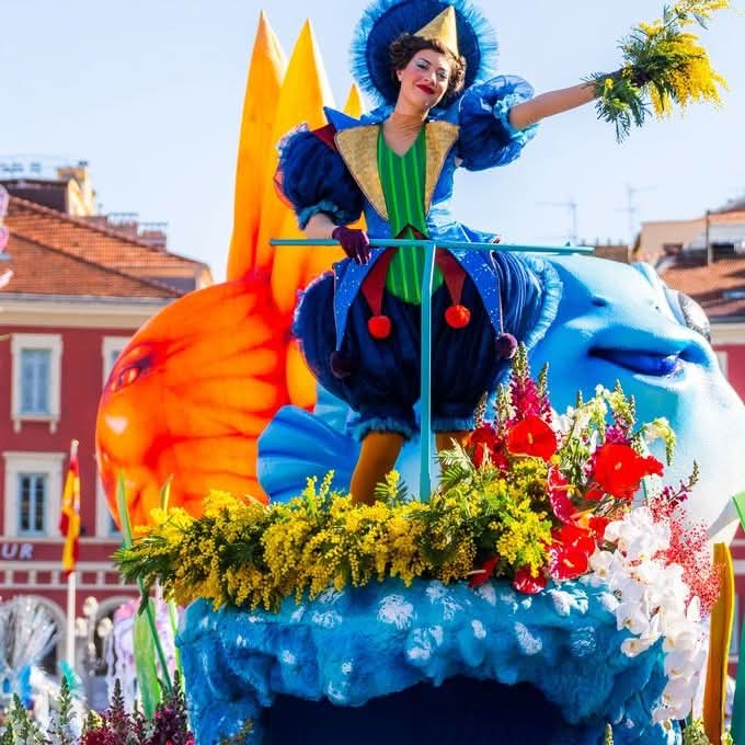 Costumed woman on a colorful carnival float with flowers and large theatrical masks