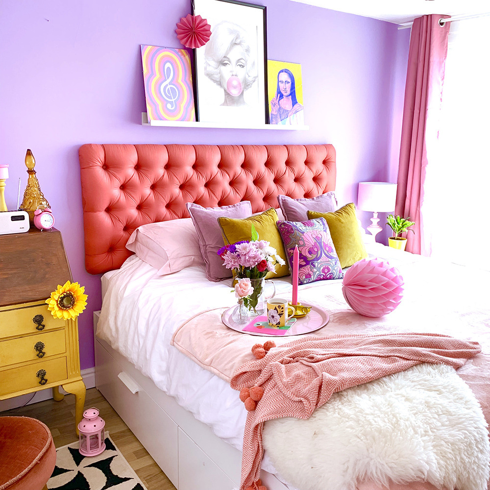 Bright bedroom with purple walls, coral headboard, white bed with pink throw, yellow nightstands, and decorative artwork