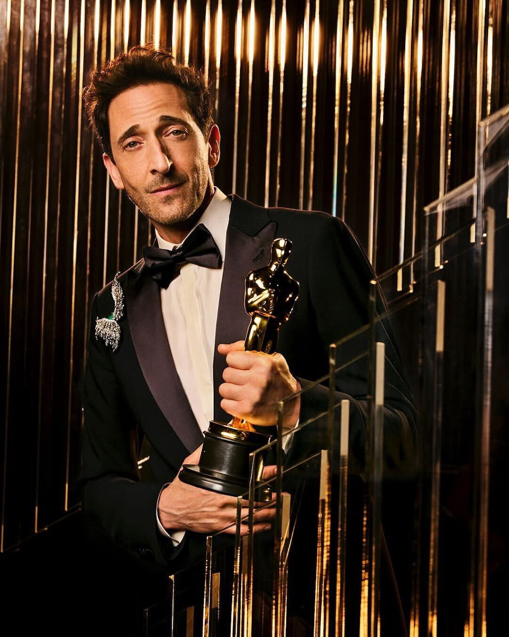 Man in tuxedo holding Oscar award statue, smiling at camera with gold striped background