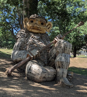 Large wooden troll sculpture in a park holding a long stringed instrument under trees
