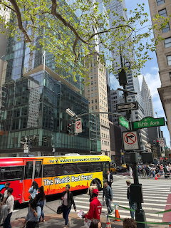 Fifth Avenue street corner in Midtown Manhattan with skyscrapers, a yellow tour bus, and pedestrians