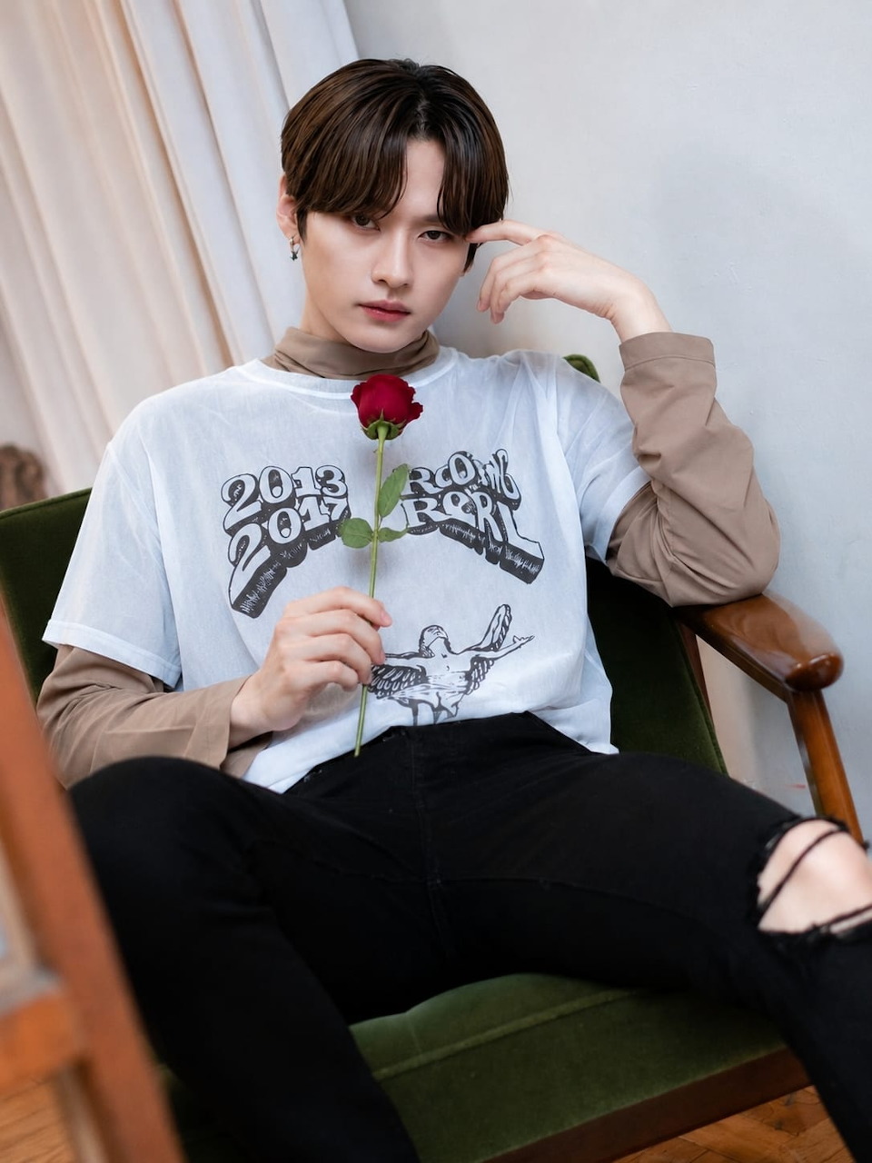 Young Asian man sitting in a green chair holding a red rose, wearing a white graphic t-shirt and black jeans