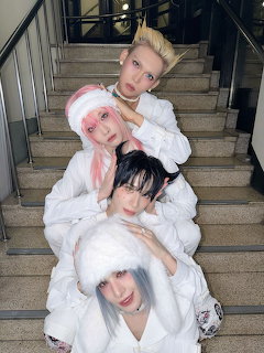 Four K-pop artists in white outfits and colorful wigs posing on stairs