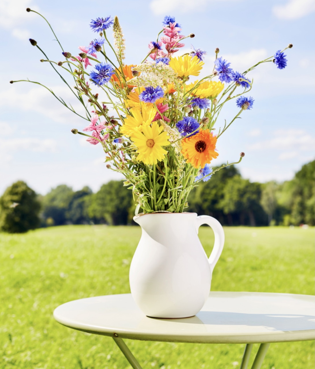 Vibrant wildflower bouquet in white pitcher on outdoor table with green lawn background
