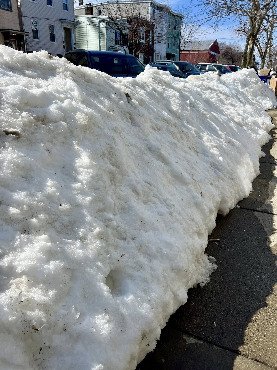 Large snow pile on a Boston sidewalk with houses and parked cars in the background