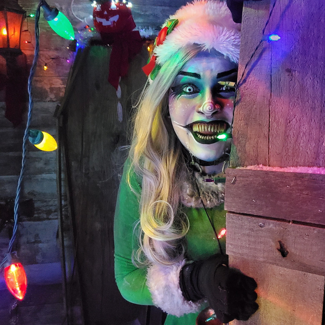 Person in Krampus helper costume with green makeup, blonde hair, and Christmas lights behind