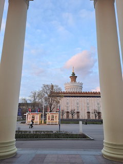 Historic ornate building viewed through white architectural columns under blue sky with bare trees
