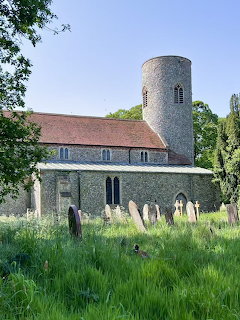 Medieval flint church with round tower and red roof surrounded by grassy graveyard with old headstones