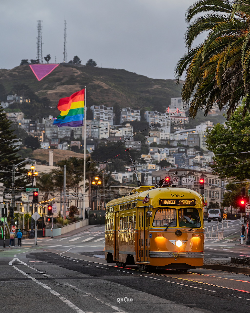 Yellow vintage streetcar on a San Francisco street with a rainbow Pride flag and hillside buildings in the background.