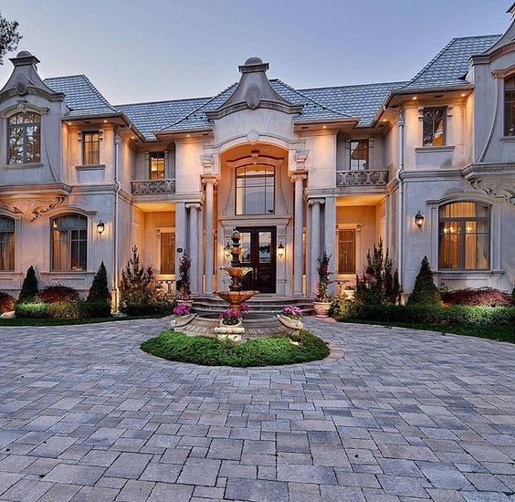 Large luxury house with lit windows and circular brick driveway at dusk