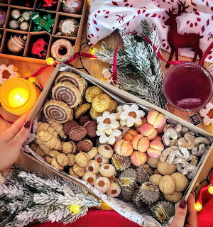 Box of assorted Christmas cookies with holiday decorations, candle, and a glass of cranberry drink