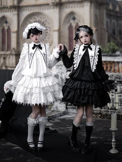 Two women in gothic lolita fashion, one in white and one in black ruffled dresses, holding hands outdoors