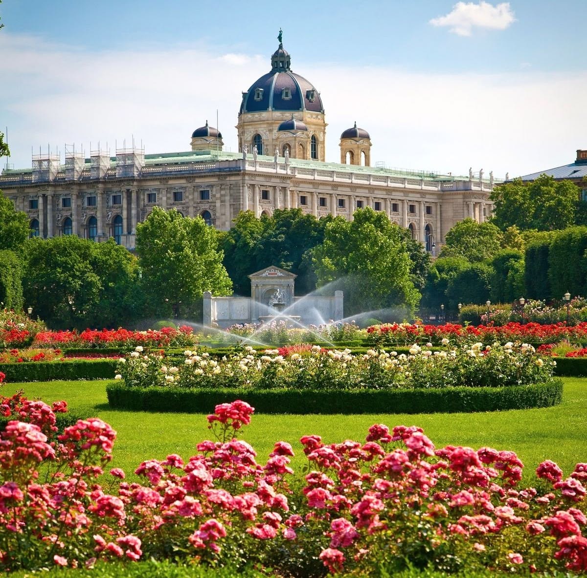 Ornate palace with dome behind formal garden beds filled with pink and red flowers, green lawn and fountain