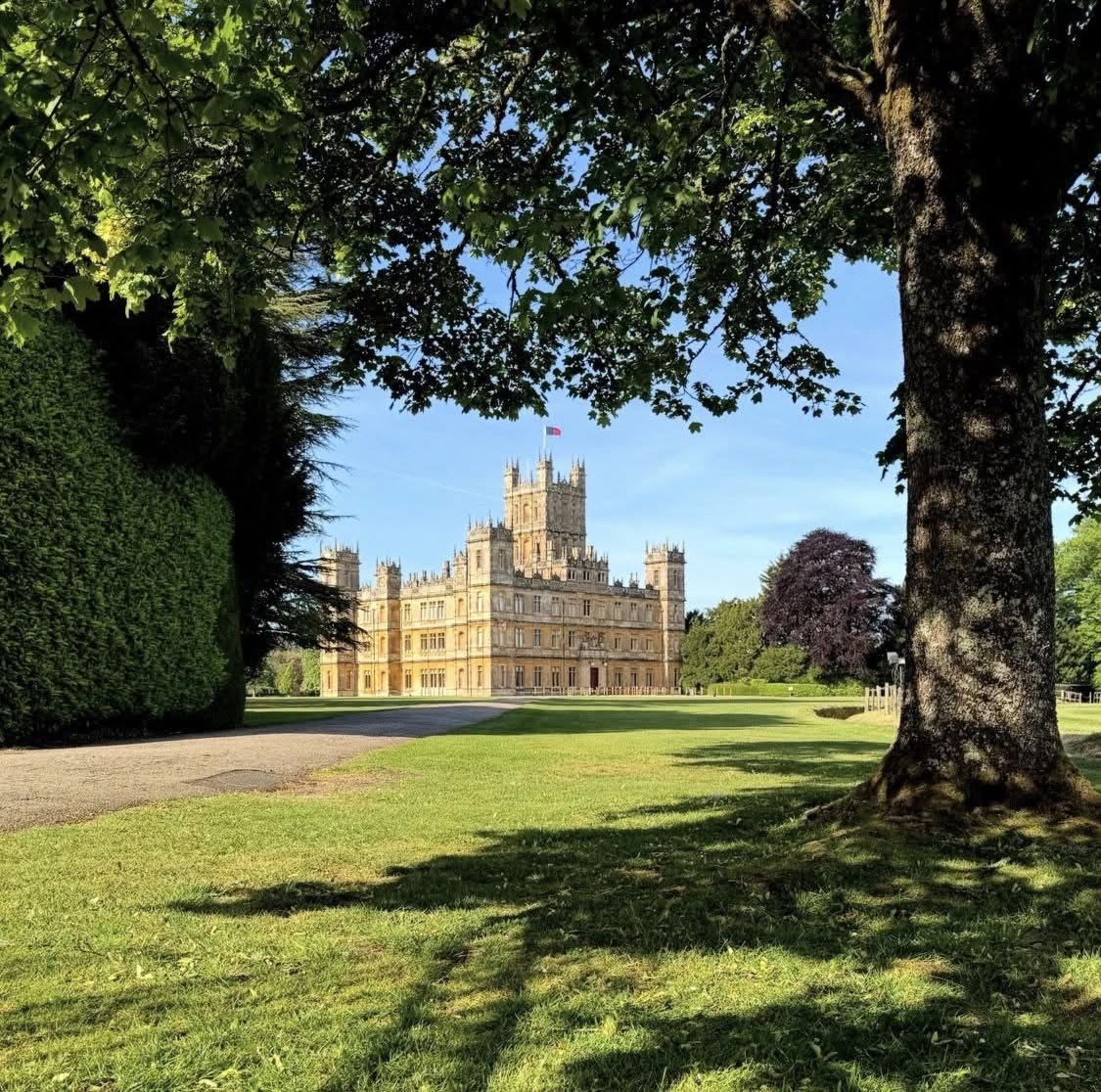 Highclere Castle viewed across a green lawn, framed by leafy trees on a sunny day