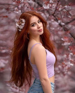 Young redhead woman in lavender top posing among pink cherry blossom branches