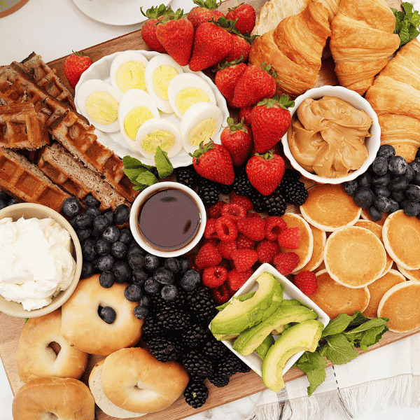 Overhead view of a breakfast spread with waffles, croissants, berries, bagels, eggs, avocado, pancakes, and coffee