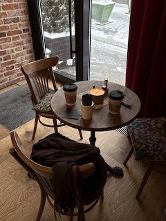 Round cafe table with coffee cups and candle near a window overlooking a snowy street, with wooden chairs.
