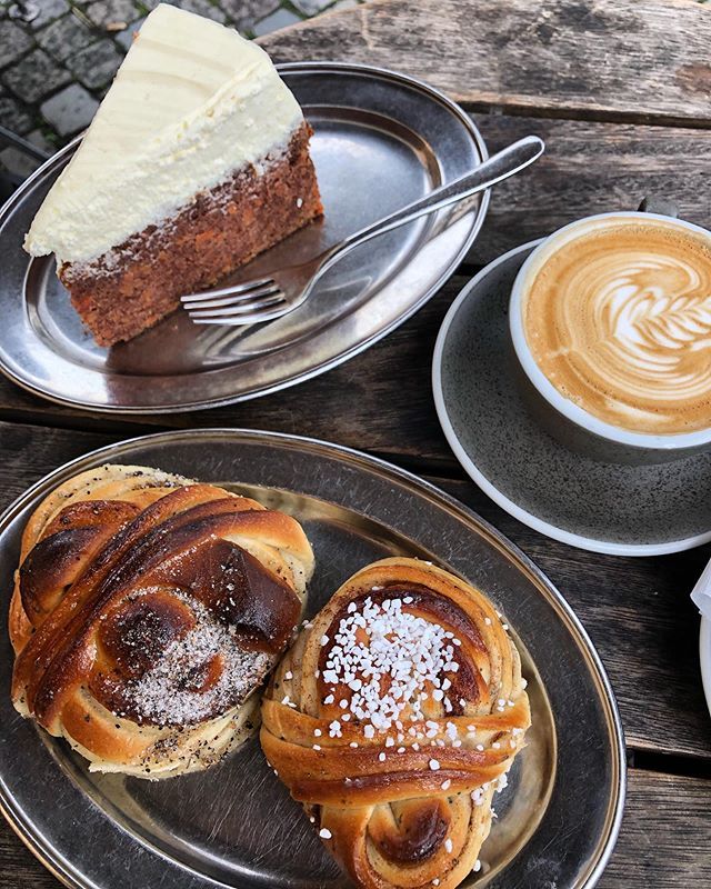 Overhead view of coffee with latte art, cinnamon buns, and carrot cake slice on dark plates over wooden table
