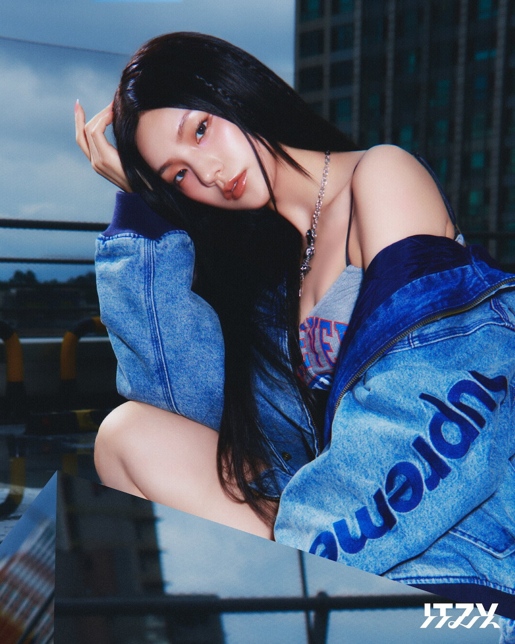 Young woman with long dark hair in denim jacket posing on roftop with city buildings in background