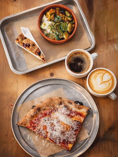 Overhead photo of lunch dishes on wooden table: pizza, salad, pie, coffee, and latte