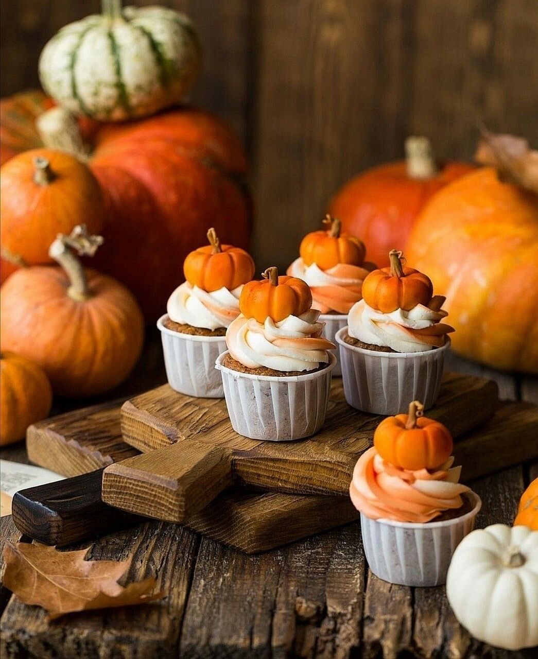 Cupcakes with cream frosting and small pumpkin decorations on wood board with fall pumpkins in background