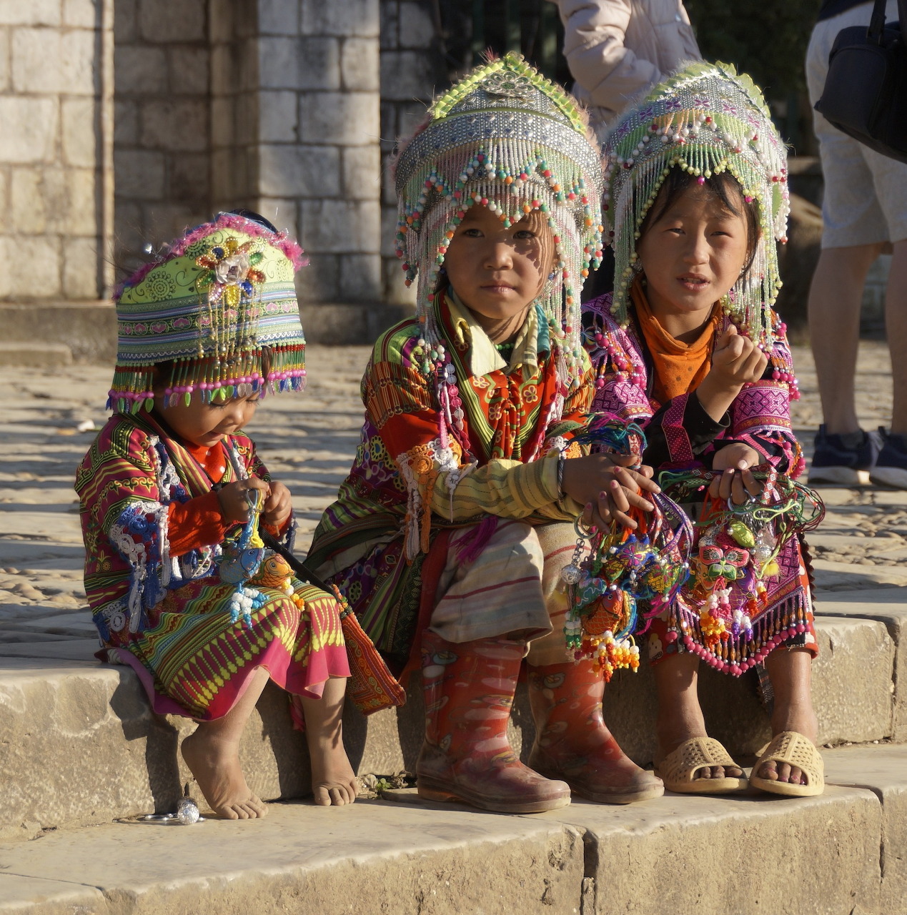 Three children in colorful traditional ethnic costumes with decorative headdresses sitting on stone steps