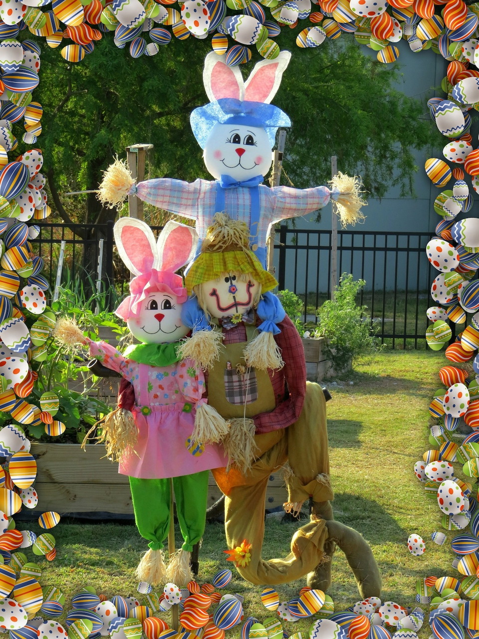 Three people in pastel bunny costumes with large heads standing on grass, surrounded by painted Easter egg frame