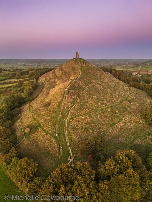 Aerial view of Glastonbury Tor at dusk with St Michael's Tower on the summit and surrounding green landscape