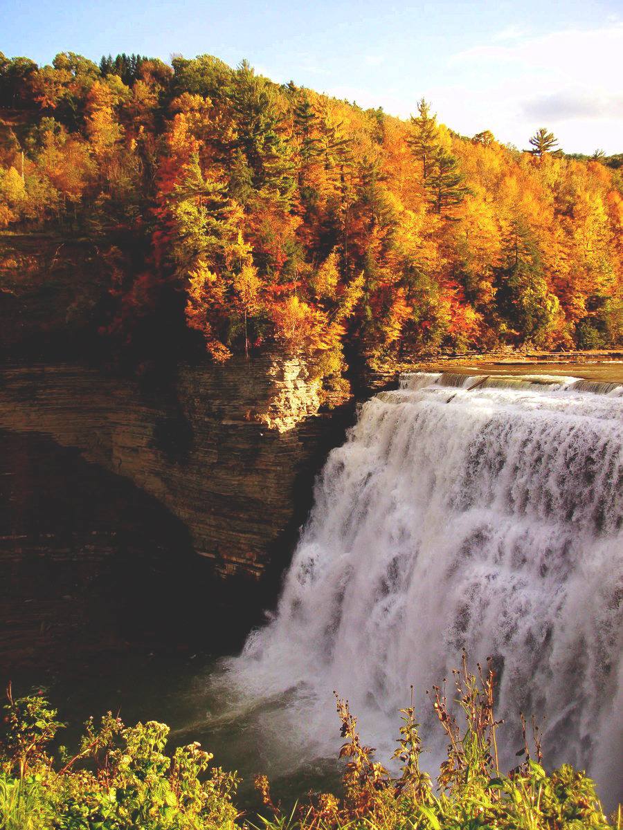 Wide waterfall flowing over layered rock cliffs surrounded by colorful autumn trees