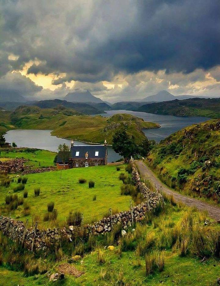 Remote stone cottage on green hillside beside loch under stormy sky in Scottish Highlands