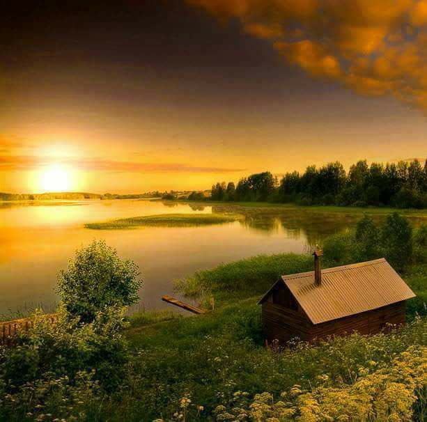 Golden sunset over calm lake with small cabin on grassy shore and dramatic orange clouds above