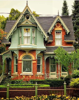Ornate Victorian house with green and orange facade, peaked gables, white decorative trim, and colorful flower garden in front