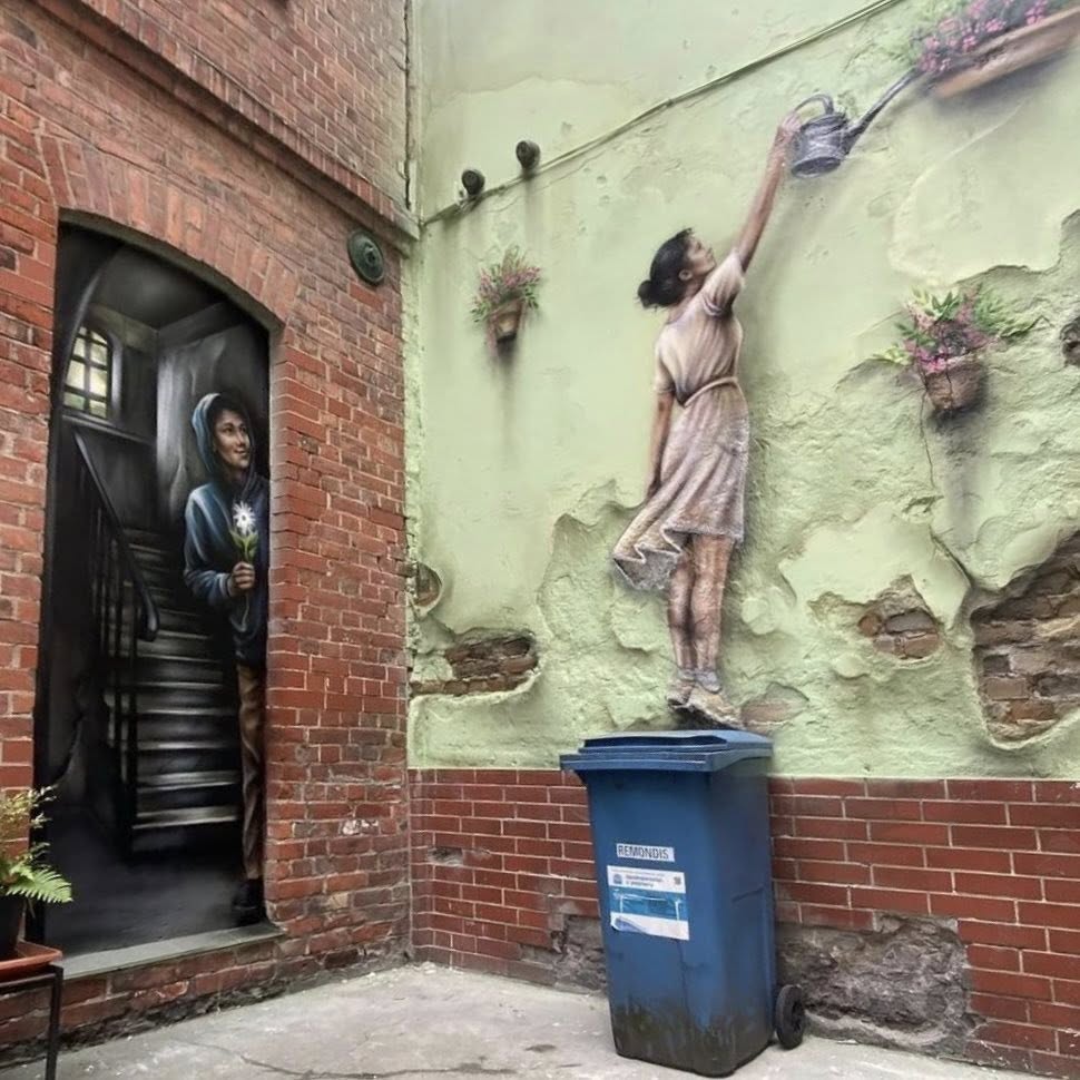 Trompe-l'oeil mural of a girl on a blue bin reaching toward flowers on an old brick wall
