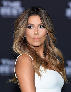 Woman with wavy brown and blonde highlighted hair wearing a white top posing against a dark background