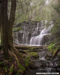 Multi-tiered waterfall cascading over rock ledges in a misty green forest with moss-covered trees