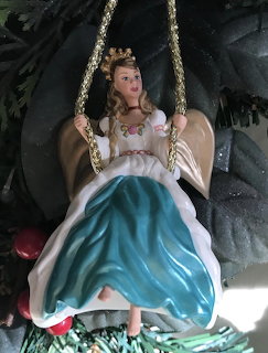 Barbie angel doll ornament on golden swing with white wings, halo, and teal dress against evergreen background