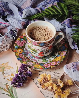 Frothy coffee in a floral patterned cup and saucer surrounded by dried flowers and vintage papers