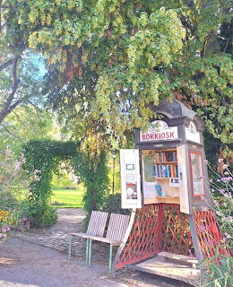 Small red and white book kiosk labeled Bokkiosk under a large tree in a green park with hedge archway