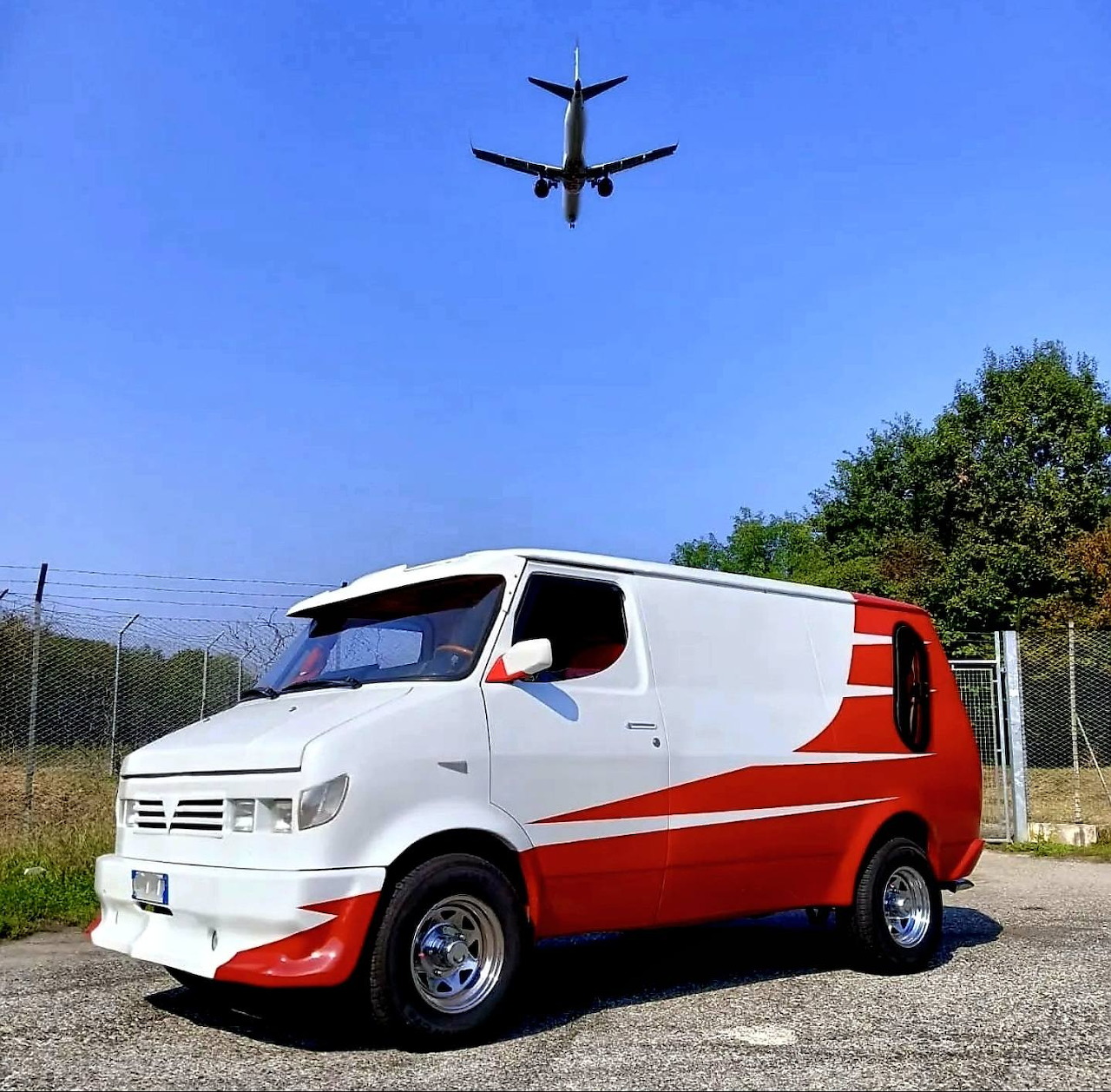 Custom red and white van parked on gravel with a plane flying overhead