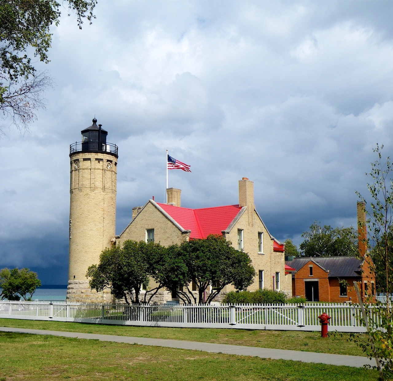 Yellow lighthouse tower with red-roofed keeper's house under cloudy sky, American flag visible, waterfront setting