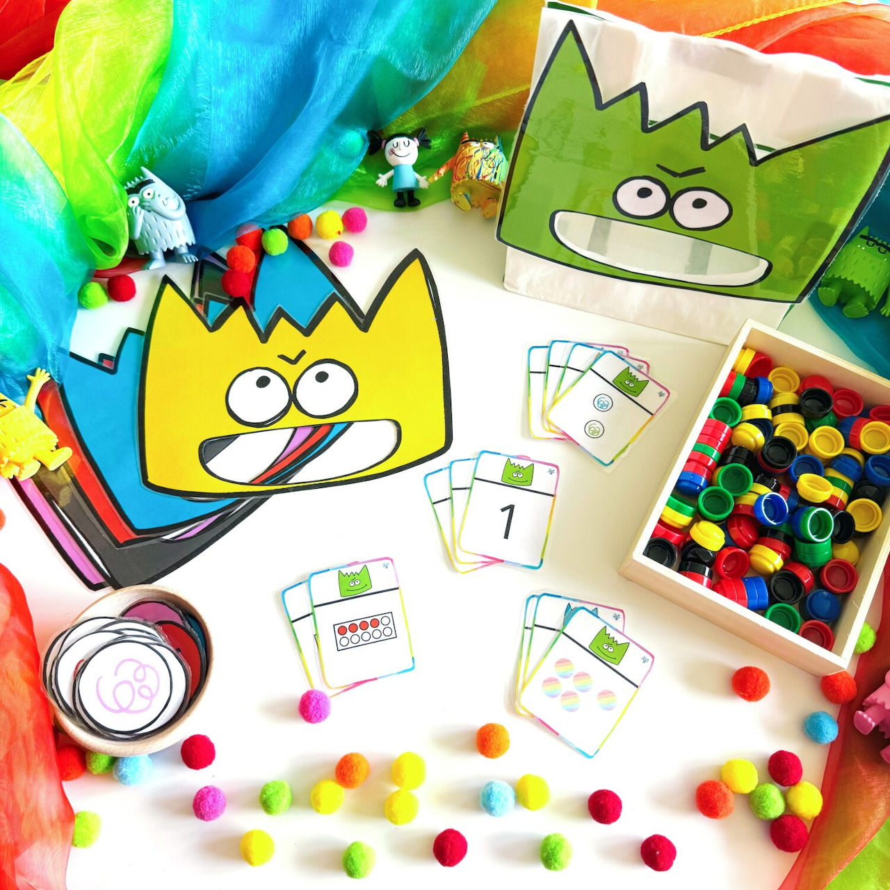Craft supplies with monster designs, colorful pom-poms, and rainbow fabric arranged on white table