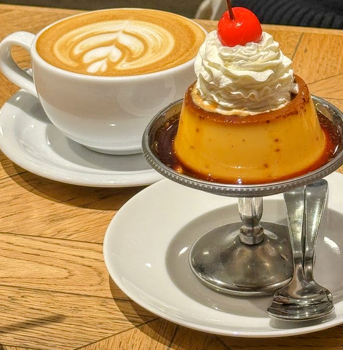 Caramel flan with whipped cream and cherry next to a cup of latte on a wooden table
