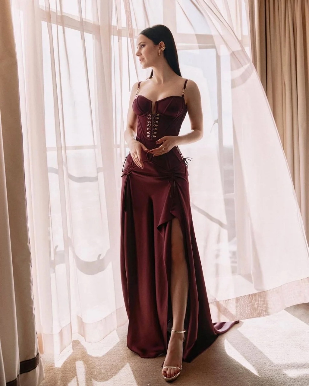 Rachel Brosnahan in a burgundy evening gown standing by sheer curtains in soft sunlight