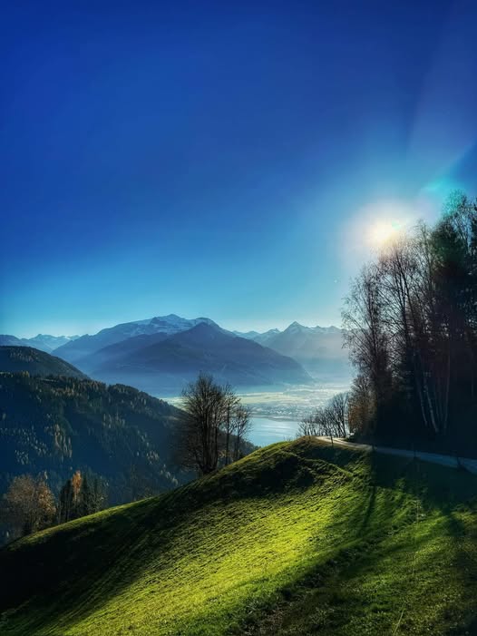 Sunlit green hills overlooking a misty Austrian alpine valley with mountains and trees under a blue sky