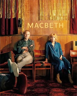 Man and woman seated in a vintage theater lobby with MACBETH text above them