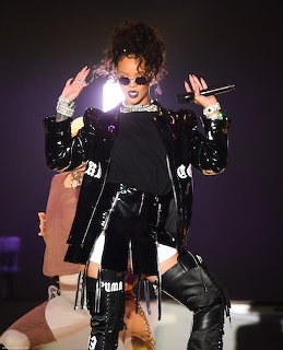 Rihanna performing on stage in black patent leather outfit with sunglasses and curly updo hairstyle