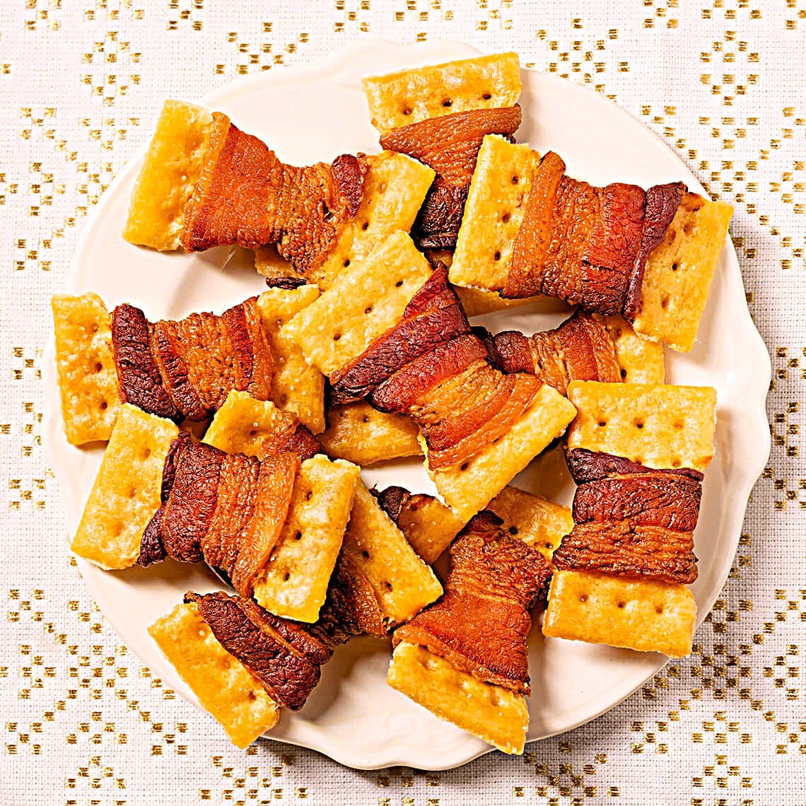 Crackers wrapped with crispy bacon on a white plate over a patterned tablecloth